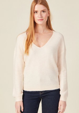 Losse crèmekleurige trui met V-hals, geribbelde textuur en lange mouwen; gecombineerd met donkere jeans, wat een casual stijl laat zien.