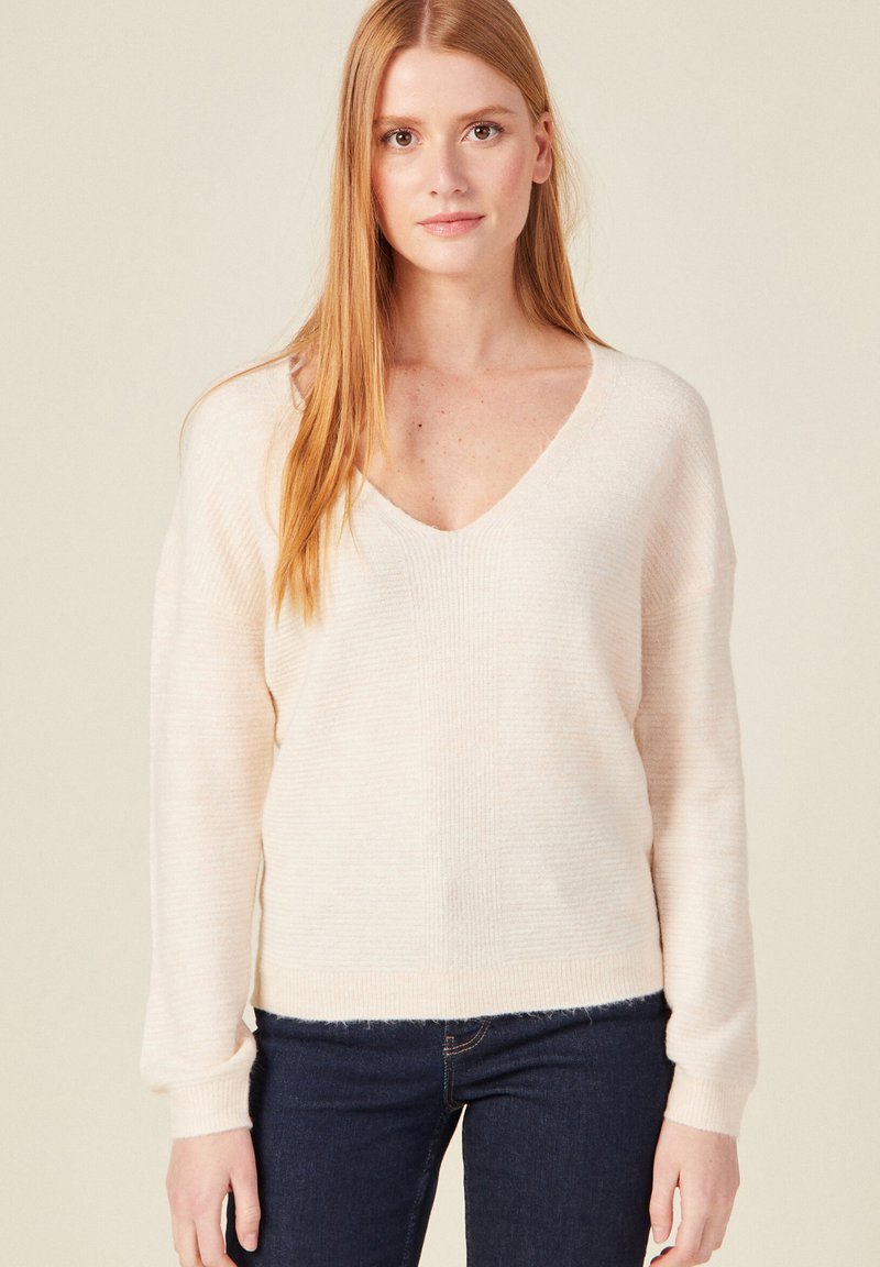 Lockere, cremefarbene Strickjacke mit V-Ausschnitt, gerippter Textur und langen Ärmeln; kombiniert mit dunklen Jeans und präsentiert einen lässigen Stil.