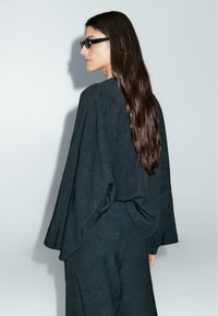 Černý, strukturovaný textilní úbor se širokými rukávy a volným střihem, s drapérií a minimalistickým stylem. Dlouhé, rovné vlasy.