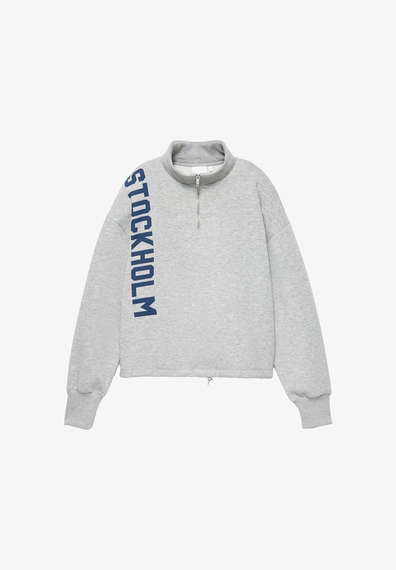 Pull gris clair à manches longues avec une courte fermeture éclair au niveau du cou et "STOCKHOLM" en grandes lettres bleues verticalement sur le côté gauche.