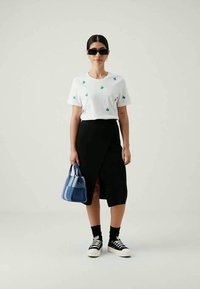 Mulher vestindo uma t-shirt branca com estampas de cactos verdes, saia midi preta com fenda frontal, sapatilhas pretas, óculos de sol e segurando uma handbag listrada azul.