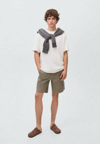 Světlý béžový tričko, šedý svetr přehozený přes ramena, olivově zelené kraťasy, hnědé nazouvací boty, stojící na jednobarevném šedém pozadí.