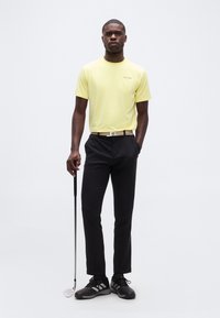 Άνδρας που στέκεται με κίτρινο πουκάμισο Calvin Klein, μαύρο παντελόνι και μαύρα αθλητικά παπούτσια, κρατώντας ένα γκολφ κλαμπ με το ένα χέρι στην τσέπη.