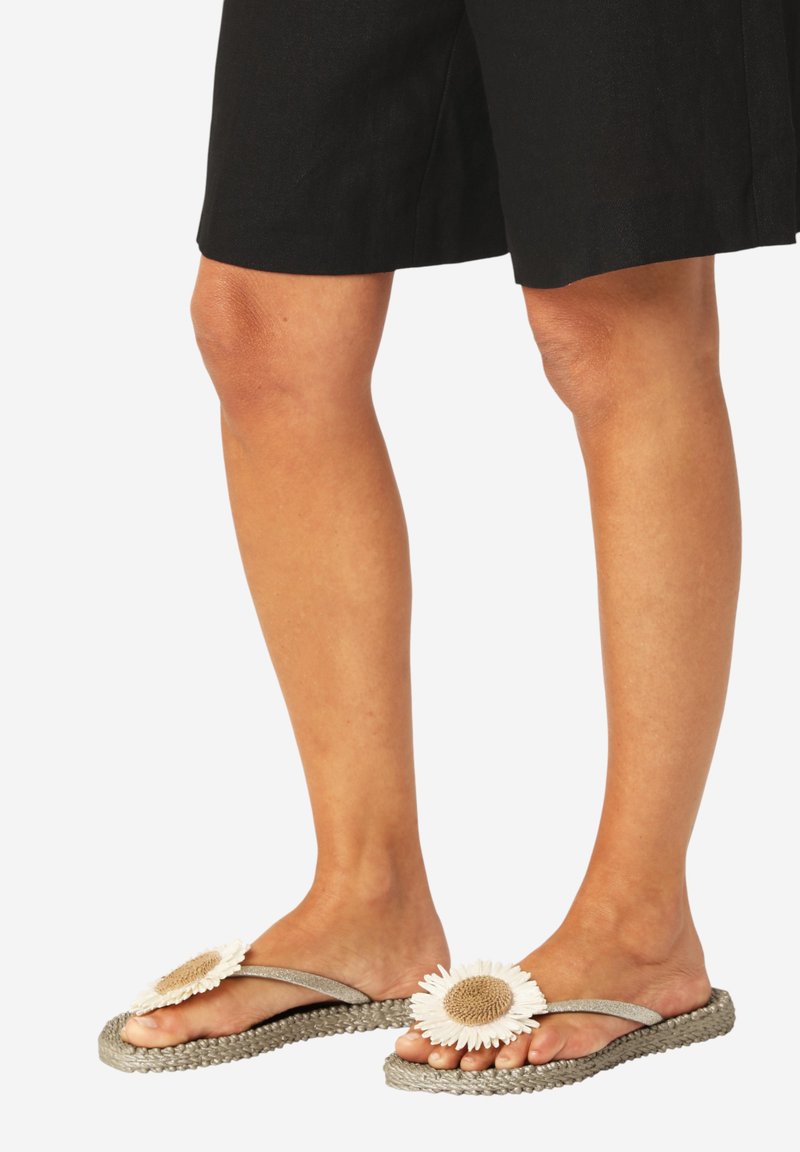 Pieds portant des sandales tissées grises avec de grandes décorations de fleurs blanches et beiges, associés à un short noir jusqu'aux genoux.