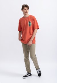 T-shirt arancione in cotone con stampa grafica sul petto, abbinata a pantaloni beige e sneakers nere. Design morbido, maniche corte.