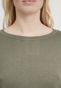 Olive zaļš ribots adījumdžemperis ar plašu piegriezuma kaklu un smalkiem šuvju detaļām, izgatavots no mīkstas auduma ar gludu tekstūru.