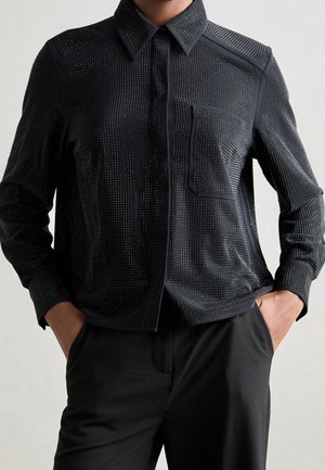 Zwart shirt met lange mouwen met kleine perforaties en een borstzak, los gedragen met een zwarte broek, handen in de zakken.