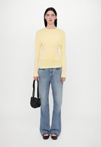 Gelbes Langarmoberteil kombiniert mit hellblauen Schlagjeans. Das Model hält eine kleine schwarze Handtasche und trägt dunkle Ballerinas.