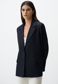Blazer blu navy con chiusura a un bottone, revers a punte e due tasche laterali. Realizzato in tessuto liscio, vestibilità sartoriale e design strutturato.