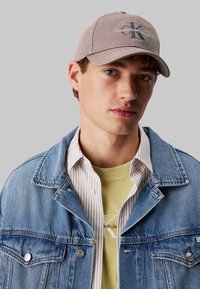 Hele hall värvi baseball müts, millel on broderitud "Calvin Klein Jeans" logo, kombineeritud sinise teksajaki ning valgete triipudega ja kollasega kihitatud särgid.