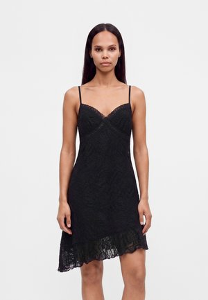 Femme aux longs cheveux foncés portant une robe noire en dentelle sans manches avec de fines bretelles et un ourlet asymétrique, debout devant un fond clair uni.