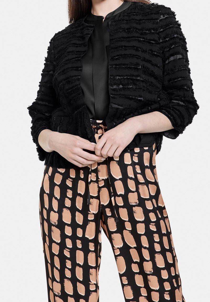 Femme portant une veste courte texturée noire, un chemisier noir et un pantalon taille haute à jambes larges avec un motif abstrait carré pêche et noir.