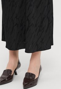 Garās pirkstu daļas, bordo krāsas lakotas ādas kurpes ar slīpu atloku dizainu; valkātas kopā ar melnu svārku ar teksturētu rakstu.