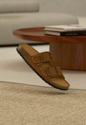 Καφέ σανδάλι με δύο ιμάντες με αγκράφες και λεπτομέρειες με τρουκς, που αιωρείται πάνω από μπεζ χαλί κοντά σε ξύλινο στρογγυλό τραπέζι με στοιβαγμένα βιβλία.