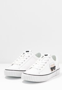 Weisse Ledersneaker mit einer strukturierten Gummisohle, schwarzem Akzentlogo und einem seitlichen Patch in neutralen Tönen. Schnürdesign mit silbernen Ösen.