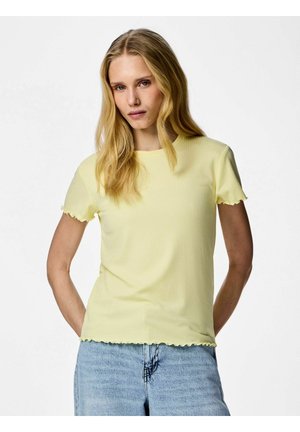 Donna bionda con capelli sciolti che indossa una maglietta giallo pallido a maniche corte con bordi ondulati e jeans in denim azzurro, in piedi davanti a uno sfondo bianco.