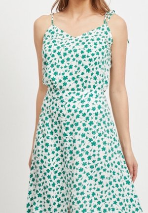 Femme portant une robe blanche sans manches avec de fines bretelles et un imprimé floral vert, debout les mains le long du corps.