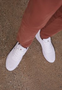 Valkoiset tennarit, joissa on teksturoitu neulepinta ja pyöreä kärki. Varustettu logotunnuksella ja nauhoilla, yhdistettynä ruosteenvärisiin housuihin betonipinnalla.