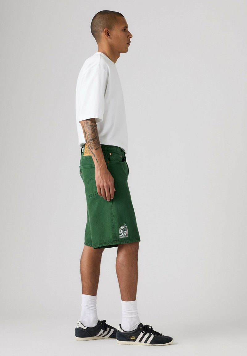 Mann trägt ein weißes Hemd, grüne knielange Shorts mit einem Mexiko-Patch, weiße Socken und schwarze Adidas-Sneaker, steht seitlich.