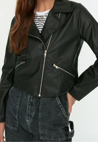 Zwarte nep leren crop jack met een geknipt kraag, een rits aan de voorkant en twee zijzakken met rits, gedragen over een gestreept shirt.