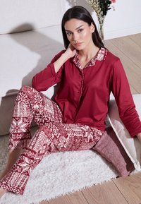 Kvinde sidder på gulvet ved sofaen iført rød knaplukket langærmet top og mønstrede røde og hvide pyjamasbukser, hviler hånden på nakken.