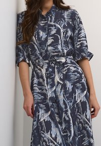 Kvinna i en marinblå knäppt klänning med vita och ljusblå abstrakta bladmönster och ett knytet midjebälte, stående mot en vit vägg.