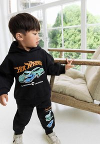 Toddler in black Hot Wheels hoodie and pants, standing indoors near wooden chair with beige cushions by a large window with greenery outside.