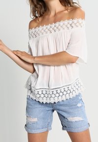 Haut blanc à épaules dénudées avec bord en dentelle, design plissé et manches courtes, assorti à un short en denim clair avec des accents déchirés.