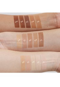 Une série d'échantillons de tons de peau sur des bras, affichant différentes nuances allant du brun foncé au beige clair, étiquetés avec des codes numériques sur un fond neutre.