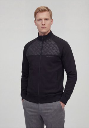 Blond man wearing a black zip-up jacket with patterned chest panel and gray pants, standing with hands in pockets against gray background.