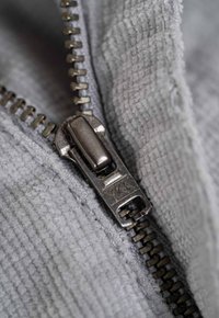 Metal zipper with a rectangular pull tab, against a soft gray corduroy fabric. Features coarse ribbed texture and YKK branding.