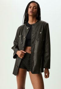 Sort faux læderjakke med en firkantet pasform, der har frontlommer, trykknapper og en glat tekstur. Bæres over en sort cropped top.