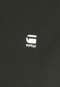 Bílé logo "G RAW" vytištěné na tmavě zelené texturované tkanině.