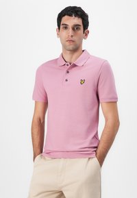 Rosa polo-skjorte med krage, tre knapper og en gul brodert logo på venstre bryst. Glatt bomullsstoff.