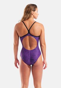 Lilla badedragt i ét stykke med tekstureret reptilmønster, åben ryg og sorte stropper, der fremhæver en slank og atletisk pasform.