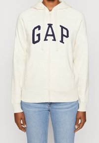 Sweat à zip crème avec logo "GAP" bleu marine, poches kangourou, capuche à cordon de serrage et poignets côtelés. Fabriqué à partir d'un matériau doux et texturé.