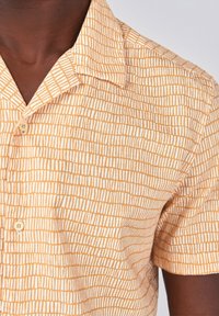 Camicia a maniche corte gialla con un motivo geometrico bianco. Presenta un colletto e dei bottoni, realizzata in tessuto leggero.