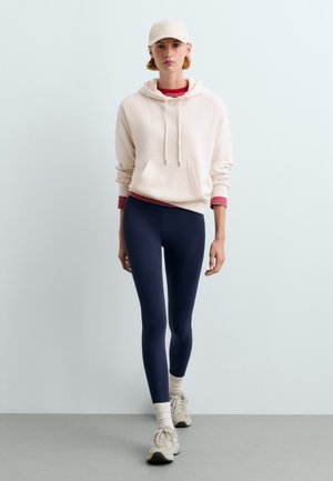 Ung person iført en hvid hættetrøje, mørkeblå leggings, hvide sneakers og en lysefarvet kasket står mod en ensfarvet lysegrå baggrund.