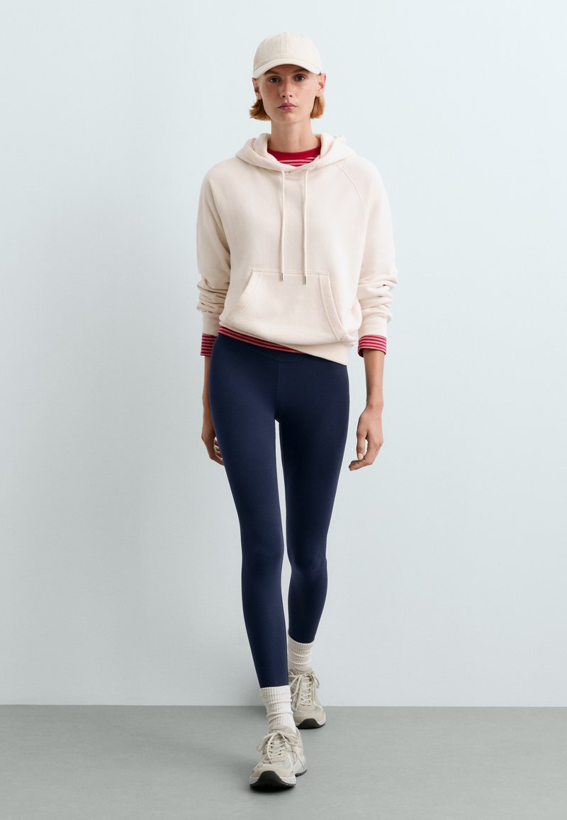Jeune personne portant un sweat à capuche blanc, des leggings bleu marine, des baskets blanches et une casquette de couleur claire, debout devant un fond gris clair uni.