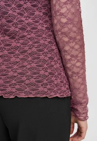 Spitzenoberteil in Mauve mit floralem Muster und langen, transparenten Ärmeln, kombiniert mit einem schwarzen Unterteil. Verfügt über einen gewellten Saum und eine zarte Textur.
