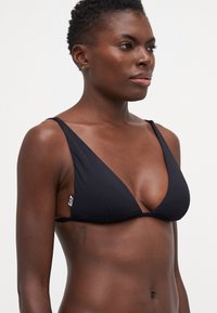 Juodas bikini viršus su giliu V formos iškirpimu, siaurais petnešėlėmis, lygia tekstūra ir mažomis baltomis logotipo detalėmis šone.