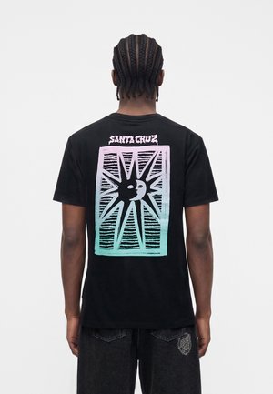 Άτομο με πλεξούδες που φοράει μαύρο T-shirt με γραφικό ήλιο και φεγγάρι σε παστέλ διαβάθμιση και κείμενο "Santa Cruz" στην πλάτη.