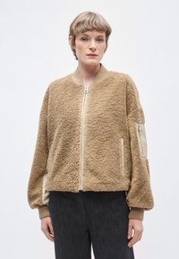 Geacă bomber căptușită, pufoasă, de culoare bronz, realizată din blană ecologică texturată cu accente bej, fermoar frontal și manșete striate. Are buzunare laterale și o cataramă la cot.