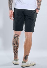 Schwarze Shorts aus Baumwolle, knielang, mit Seitentaschen. Auffällige Tattoos an den Beinen und weiße Sneakers mit grünen Akzenten.