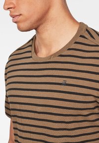 Camiseta de rayas marrón con rayas horizontales negras, cuello redondo, textura suave y un pequeño detalle de logo en el pecho.