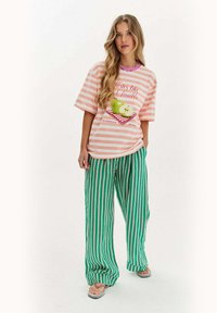 Mladá žena s dlhými vlasmi, oblečená v prúžkovanej ružovej a bielej košeli s grafickým motívom jablka, širokých nohaviciach s prúžkami v zelenej a bielej farbe a sandáloch.
