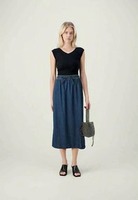 Top preto ajustado, saia maxi de denim com cintura elástica e uma bolsa preta texturizada com pega. A modelo usa sapatos pretos de salto baixo.