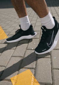 Jambes portant des chaussettes blanches et des baskets noires debout sur un trottoir gris avec des flèches jaunes.