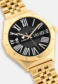 Gold-tone metal watch with a squared case, black dial, bold white Roman numerals, and textured accents. Features heart rate and step count display.