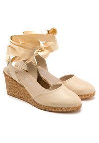 Beżowe sandały na koturnie z skórzaną cholewką, otwartym noskiem oraz dekoracyjną satynową wstążką owiniętą wokół kostki. Teksturowana plecionka w śródstopiu.